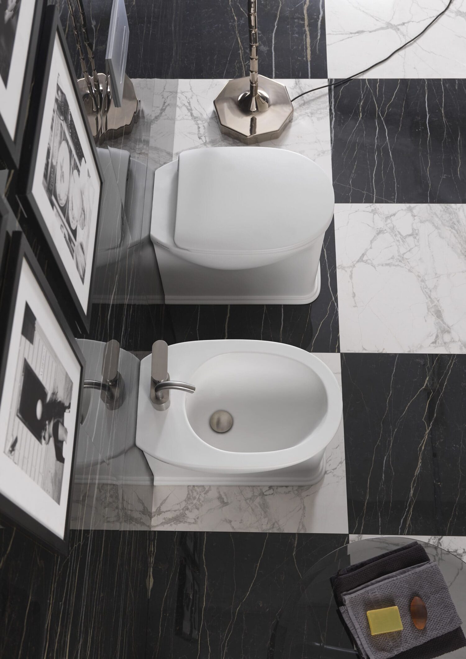 Modern bathroom features a toilet, bidet, and black-and-white checkered marble floor. Framed photos adorn the wall; a towel and soap rest nearby.