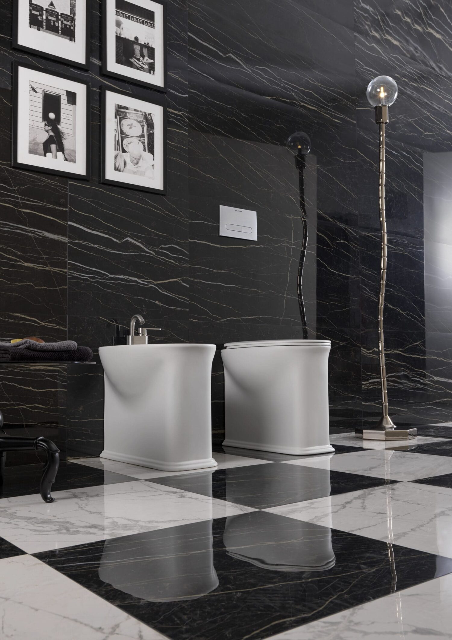 Luxurious bathroom with black marble walls, checkerboard floor, modern fixtures, and framed photographs. Elegant lamp adds stylish ambiance to the space.