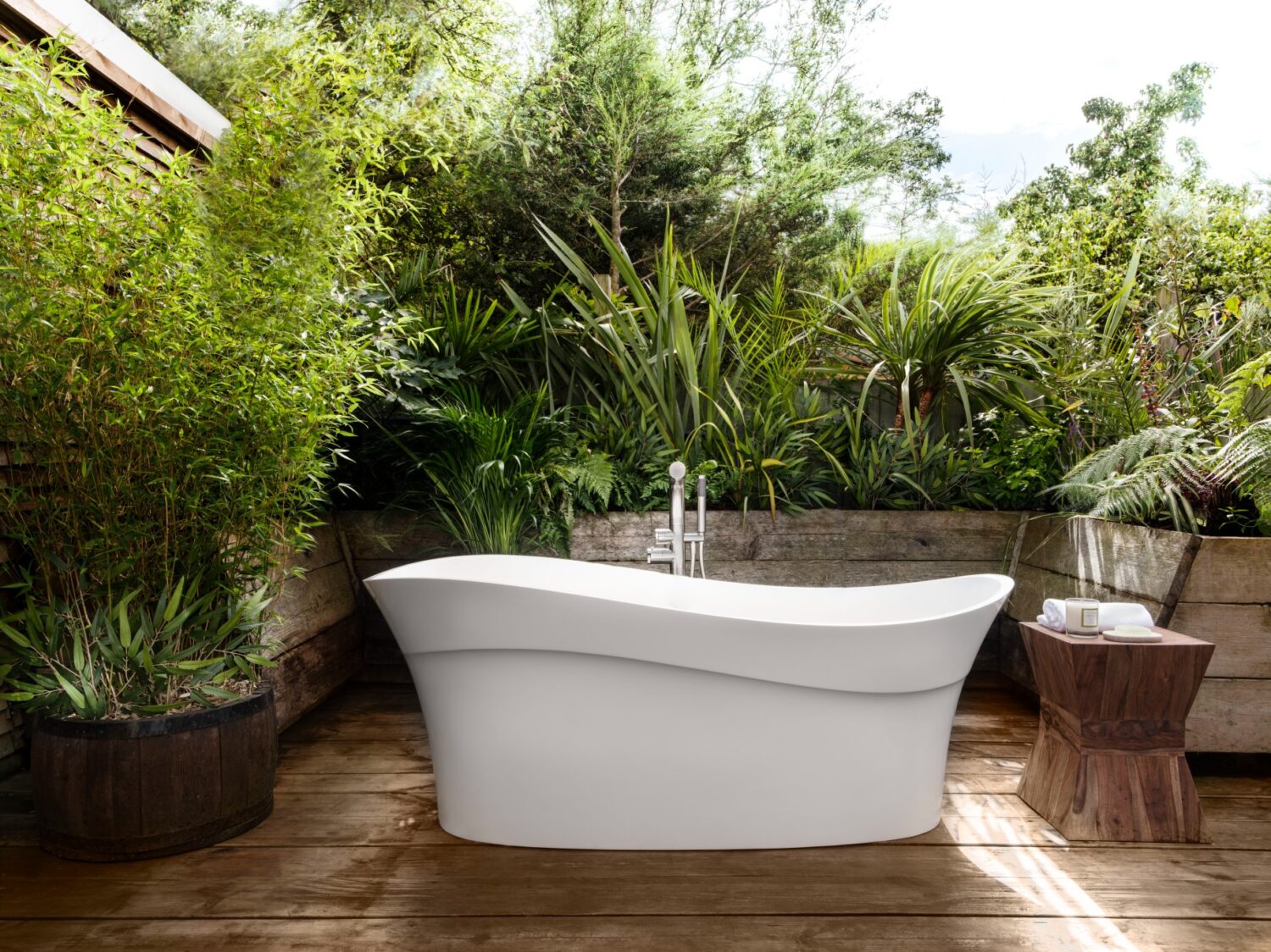 Outdoor spa area with a white bathtub surrounded by lush green plants, wooden decking, and a small rustic side table. Peaceful atmosphere.