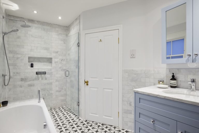 Modern bathroom with marble tiles, glass shower, bath, and blue vanity. Patterned floor tiles add contrast in a sleek, minimalist design.