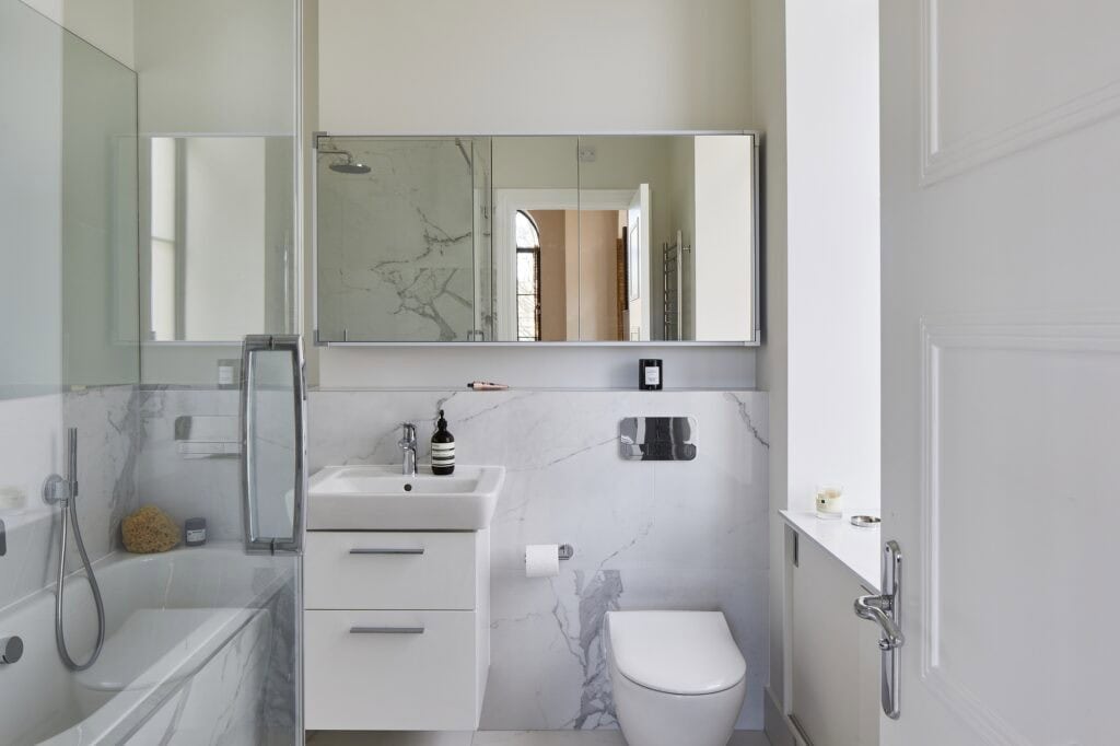 Modern bathroom with marble accents, featuring a bathtub with shower, sink with mirror above, and toilet. Minimalist design, natural light through window.