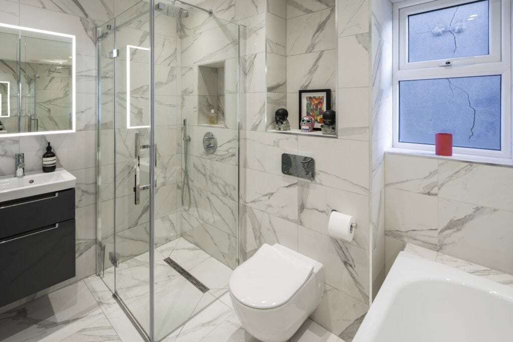 Modern bathroom with marble tiles, glass shower enclosure, and sleek fixtures. Decor includes colorful artwork and skull figurines. Window shows exterior cracks.