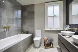 Modern bathroom features a bathtub, toilet, and sink with gold fixtures. Grey tiles and neutral colors create a sleek, minimalist design.