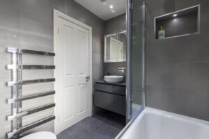 Modern bathroom with gray tiles, glass shower door, white basin, and sleek metal towel rack. Minimalist design with built-in shelf lighting.