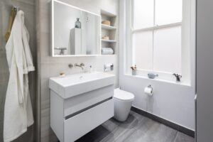 Modern bathroom with white fixtures, illuminated mirror, and gray tiles. Features a neatly organized shelf, large window, and hanging robe next to the sink.