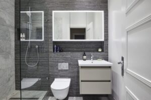 Modern bathroom features a glass shower, sleek mirror with backlighting, contemporary sink, and grey tiled walls, creating a clean and minimalist design.