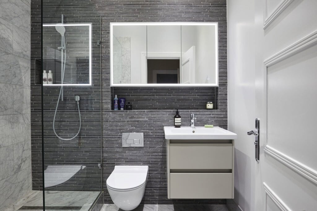 Modern bathroom features a glass shower, sleek mirror with backlighting, contemporary sink, and grey tiled walls, creating a clean and minimalist design.