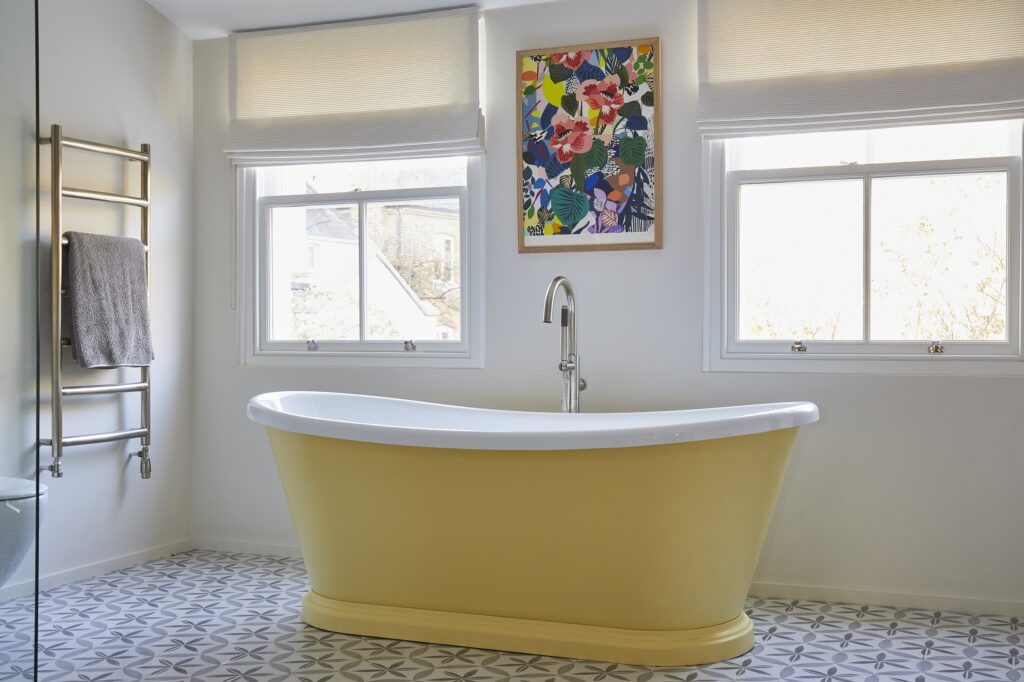 Bright bathroom with yellow bathtub, geometric floor tiles, and vibrant floral artwork. Two windows provide natural light; a towel hangs on a heated rack.