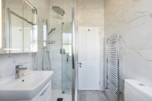 A modern bathroom featuring a glass shower enclosure, sleek fixtures, a white sink, a towel rack, and marble walls, offering a clean aesthetic.