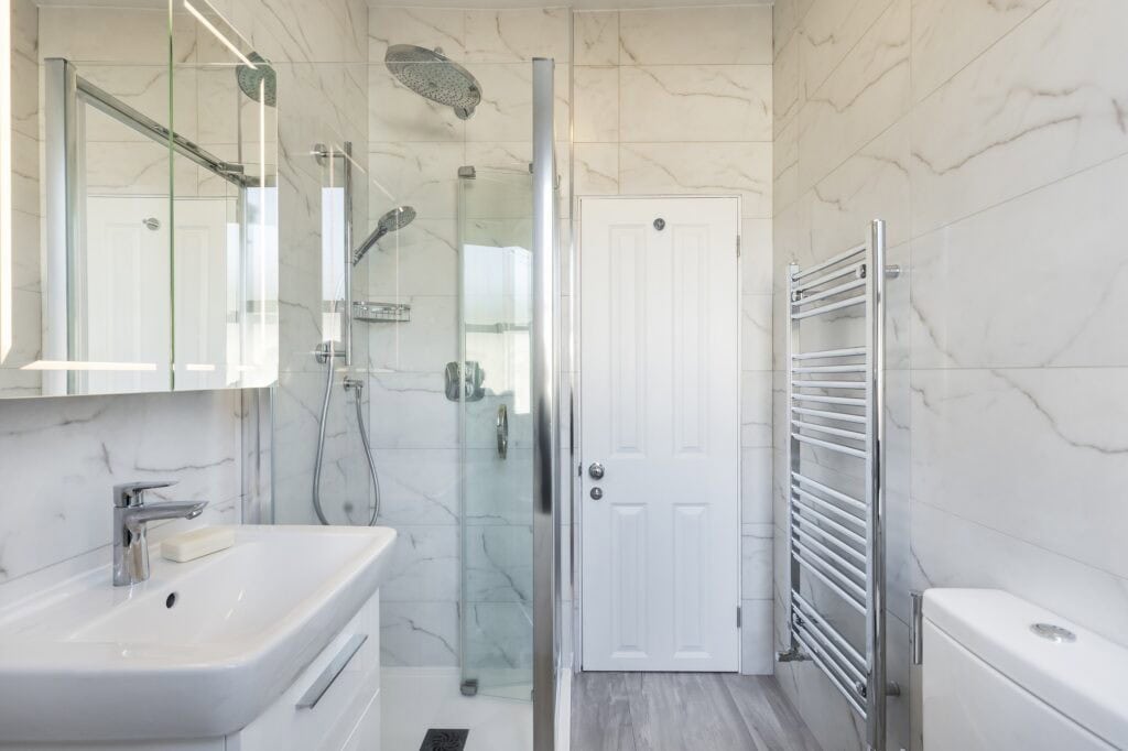 A modern bathroom featuring a glass shower enclosure, sleek fixtures, a white sink, a towel rack, and marble walls, offering a clean aesthetic.