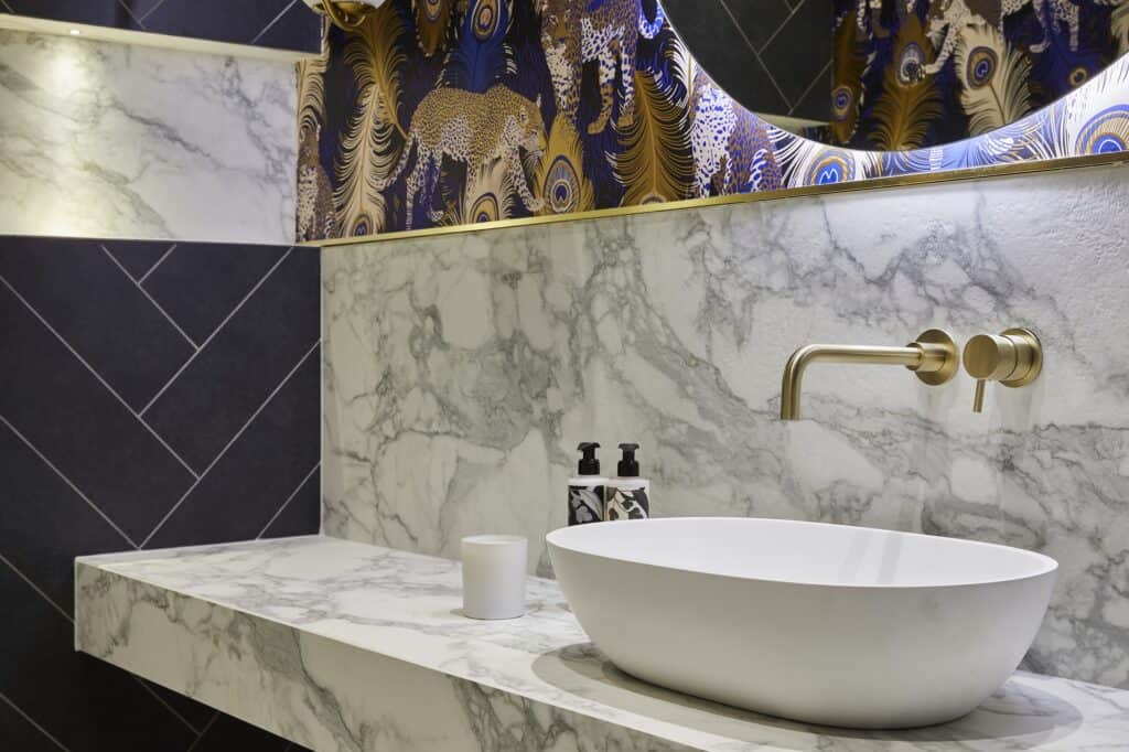 Stylish bathroom with a marble countertop, brass faucet, decorative mirror, and patterned wallpaper featuring peacock feathers and leopards. Modern and elegant design.