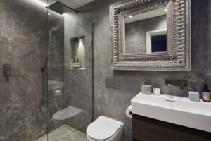 A modern bathroom with gray marble walls, a large ornate mirror, glass shower enclosure, and sleek white fixtures. Luxurious and minimalist design.