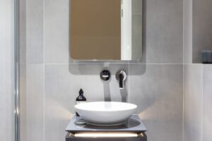 Bathroom interior with modern design features a round sink, wall-mounted faucet, square mirror, and toiletries on a sleek countertop with gray tiles.