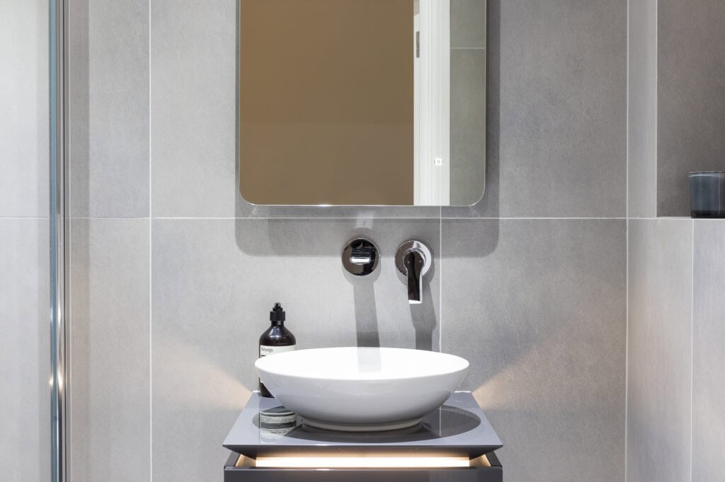 Bathroom interior with modern design features a round sink, wall-mounted faucet, square mirror, and toiletries on a sleek countertop with gray tiles.