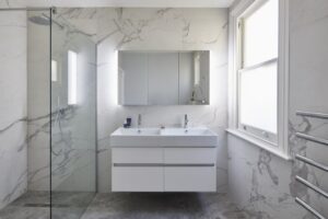 Modern bathroom with marble walls, glass shower, large mirror, and white sink cabinet. Natural light enters through a window, creating a bright atmosphere.