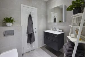 Modern bathroom with light gray tiles, wall-mounted sink, and shelf. White door with robe, large mirror, potted plants, towels neatly arranged.