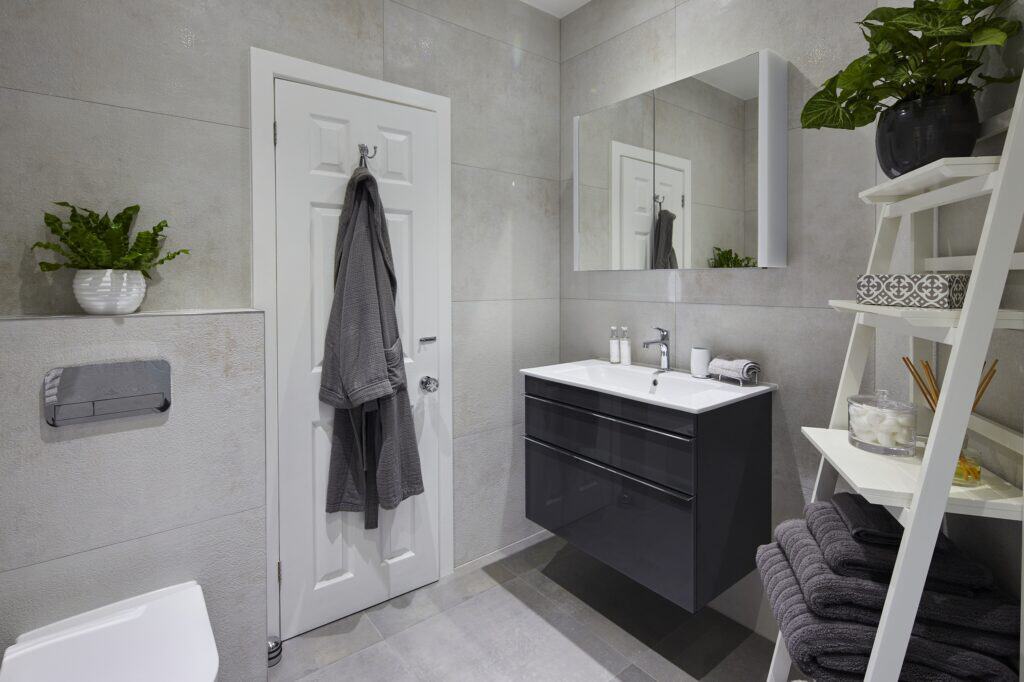 Modern bathroom with light gray tiles, wall-mounted sink, and shelf. White door with robe, large mirror, potted plants, towels neatly arranged.
