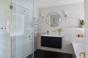 Modern bathroom with marble walls, glass shower enclosure, black vanity, circular mirror, and potted plant. Bottles and flowers decorate the countertop.