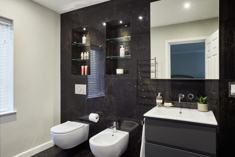Modern bathroom with black marble walls, floating toilet, and bidet. Glass shelves hold toiletries. Large mirror above sleek sink with decorative plant.