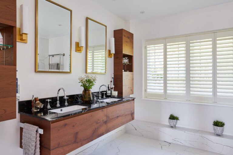 Modern bathroom with dual sinks, wood accents, and marble counter. Double mirrors and white shutters enhance the spacious, minimalist design. Plants add decoration.