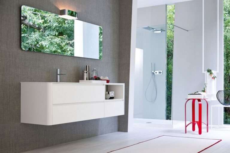Modern bathroom with white vanity, large mirror, and glass shower. Red stool holds decorations. Wall and floor feature simple, elegant design with greenery outside.