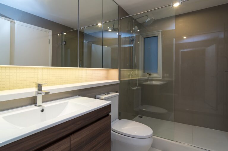 A modern bathroom with a large mirror, shower, and sleek vanity. The room has neutral tones and subtle lighting.
