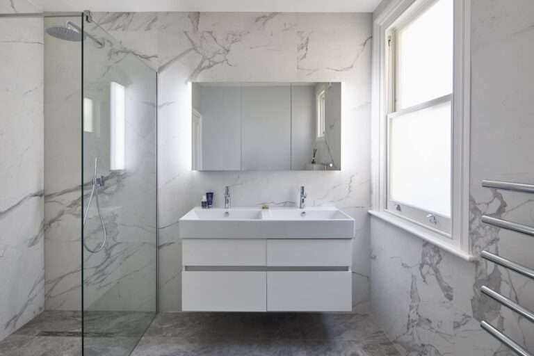 Modern bathroom with marble walls, glass shower, floating vanity, and large mirror. Bright natural light floods through the window, enhancing the minimalist design.
