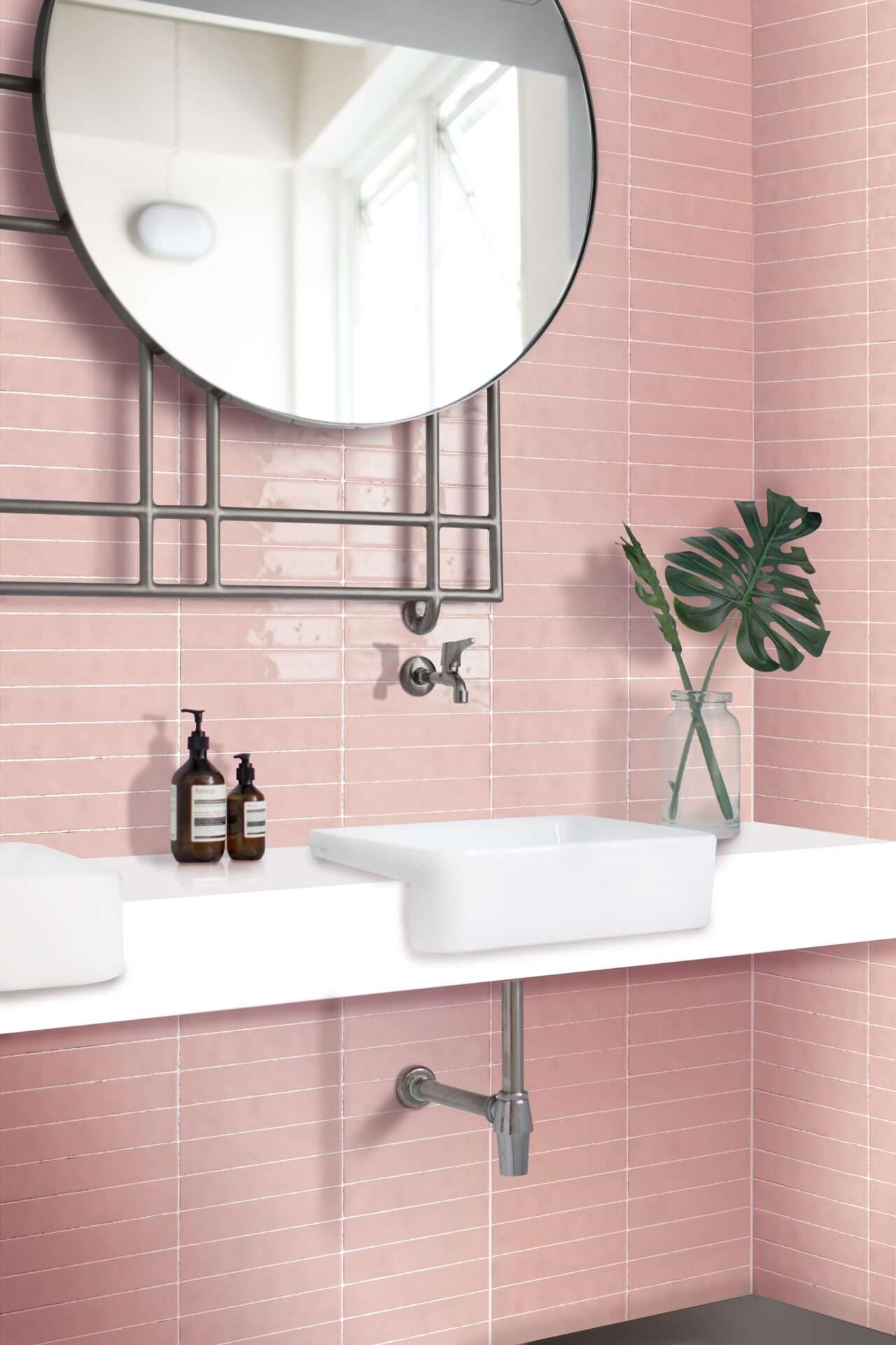 Stylish bathroom with pink tiled walls, round mirror, modern sink, and green leaf in glass vase. Two soap dispensers sit on countertop.