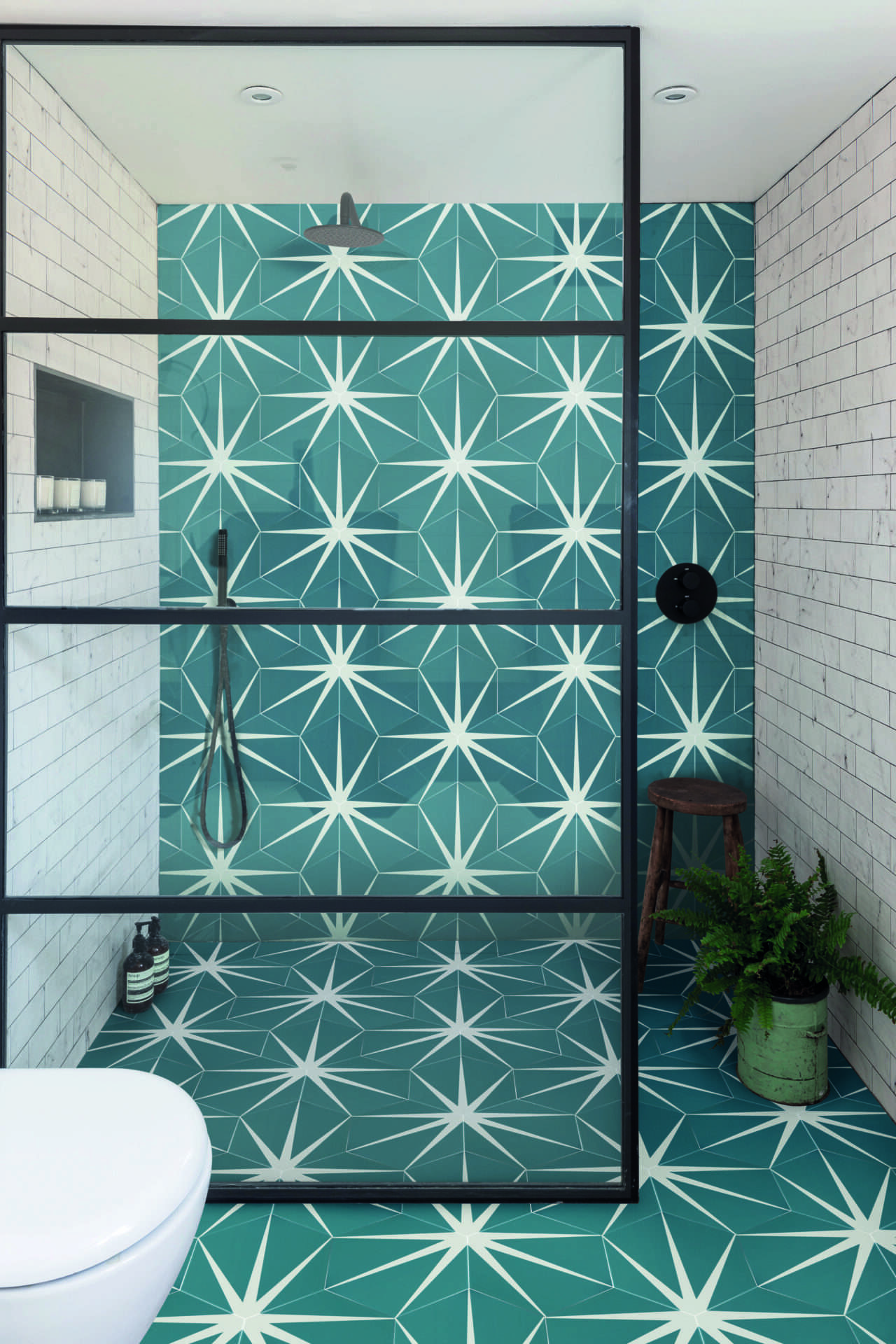 Modern bathroom featuring geometric teal and white tiles, glass shower enclosure, wooden stool, potted fern, and white brick walls. Minimalist design style.