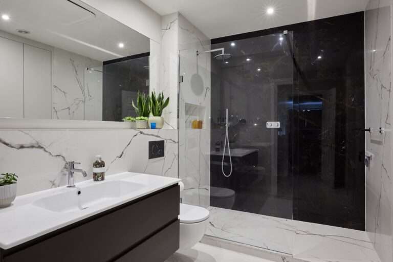 Modern bathroom with marble walls, glass shower enclosure, and sleek fixtures. Potted plants add greenery, while dim lighting creates a cozy ambiance.