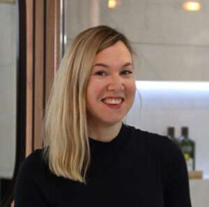 A person with blond hair is smiling indoors. The background shows shelves with bottles and soft lighting, creating a warm atmosphere.