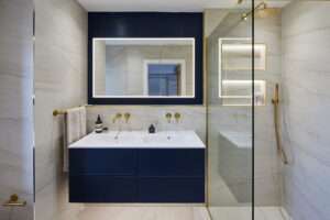 Modern bathroom with a sleek navy vanity, illuminated mirror, and gold fixtures. Marble walls and a glass shower enhance the contemporary design.