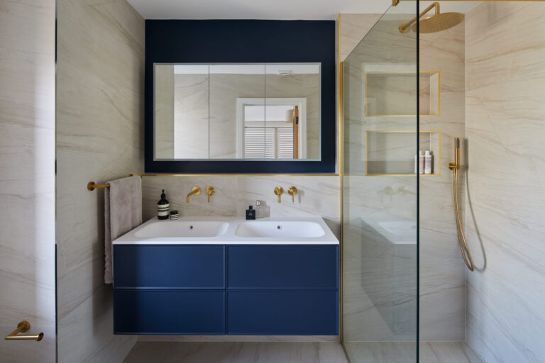 Modern bathroom with double sink, navy cabinetry, large mirror, glass shower enclosure, gold fixtures, and cream walls. Stylish design creates a luxurious atmosphere.
