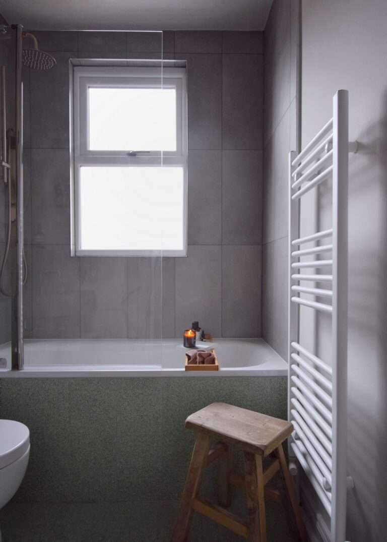 A minimalist bathroom with a window, bathtub, and wooden stool. Candles and toiletries on the tub's edge create a serene atmosphere.