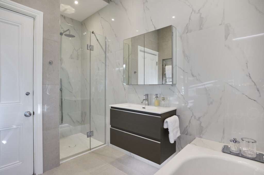 Modern bathroom with a glass shower, marble walls, and a sleek vanity. Features reflective mirror cabinet and towel hanging by the sink.