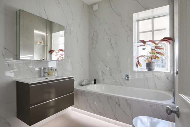 A modern bathroom with marble walls, a sleek floating vanity, a bathtub, and a potted plant on the windowsill.