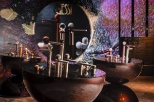 Modern art installation featuring sleek, metallic designs and spherical shapes. Vibrant background with abstract patterns. Labels reading "Gessi" visible in the scene.