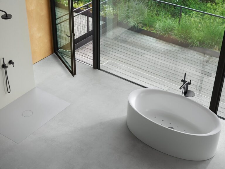 Modern bathroom with oval bathtub, sleek shower, and large glass doors opening to a wooden deck with lush greenery outside. Minimalist design and neutral tones.