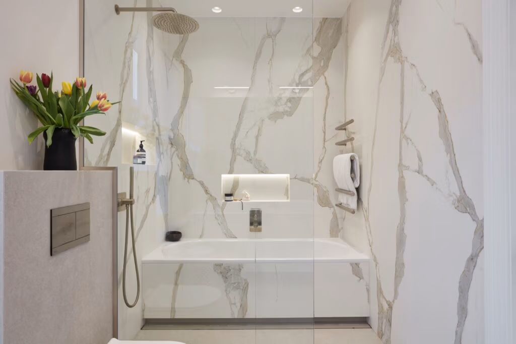 Luxurious bathroom with white marble walls, featuring a bathtub and glass shower. Tulips in a vase add color, while soft lighting creates a serene ambiance.