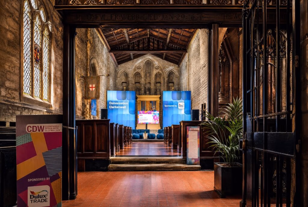 Interior of a historical church setting with a modern event setup. Blue banners display "Conversation at Clerkenwell," flanking a central screen.