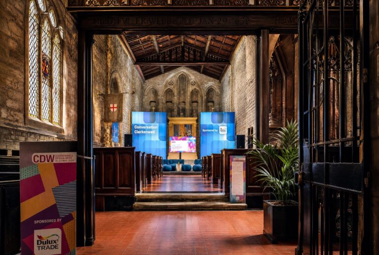 Interior of a historical church setting with a modern event setup. Blue banners display "Conversation at Clerkenwell," flanking a central screen.