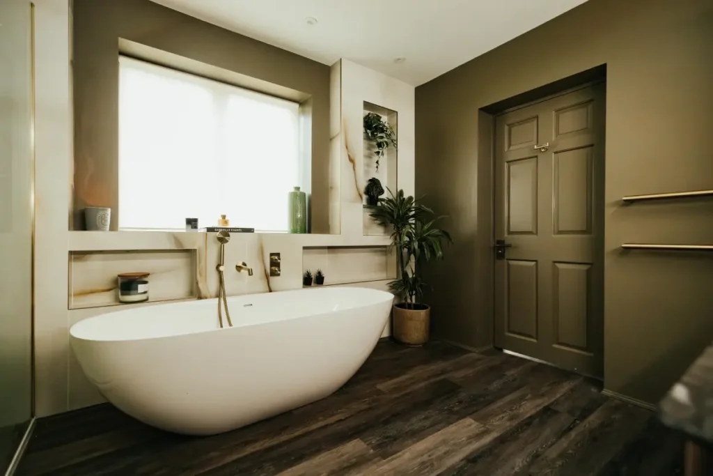 Freestanding white bath in an olive green bathroom with brushed brass fixtures, marble-effect feature wall and dark wood-effect flooring.