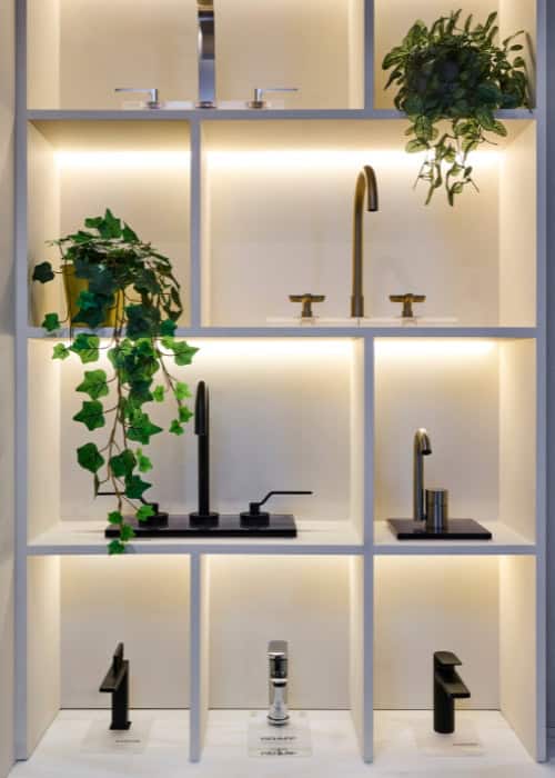 Modern bathroom fixtures with black and gold taps on illuminated shelves with plants.