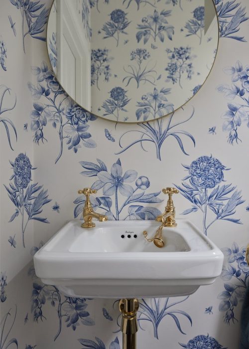 Bathroom sink with gold taps and a round mirror, decorated with blue floral wallpaper.