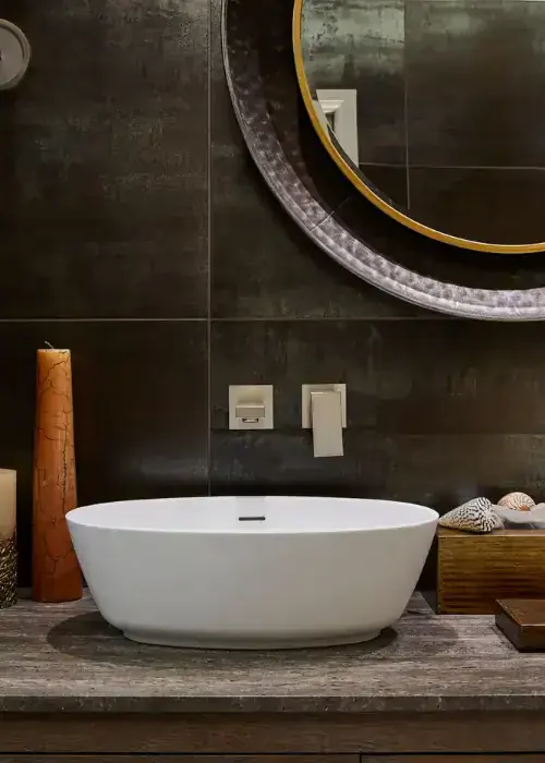 Modern freestanding bathtub in a stylish bathroom with dark tiles and round mirror.