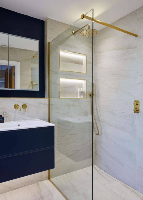 Modern bathroom features glass shower, gold fixtures, and sleek navy vanity against marble walls. LED-lit shelves add a contemporary touch.