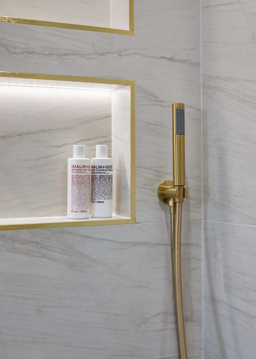 A modern shower with gold fixtures and a niche holding two bottles of Malin+Goetz products against marble-patterned tiles.