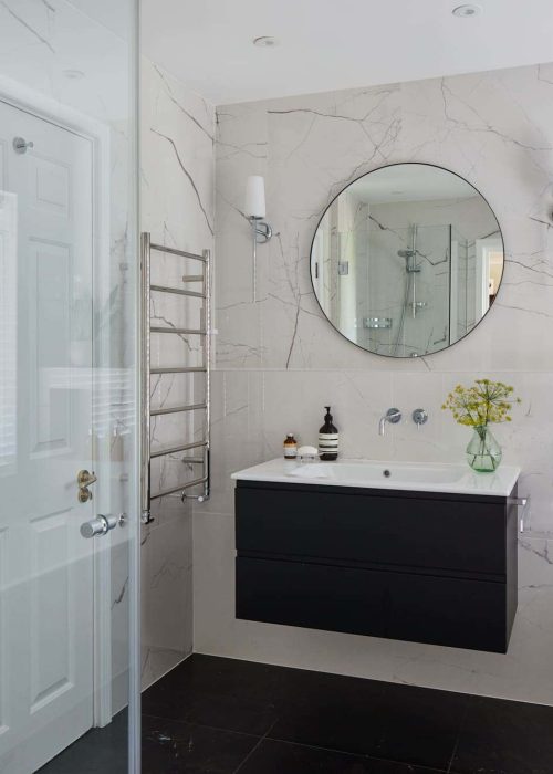 Modern bathroom with marble walls, glass shower enclosure, black vanity, circular mirror, and potted plant. Bottles and flowers decorate the countertop.