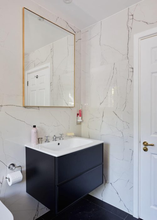 Bathroom interior features a large mirror, marble walls, white sink with black cabinet, and a door with a gold handle. Minimalist design.