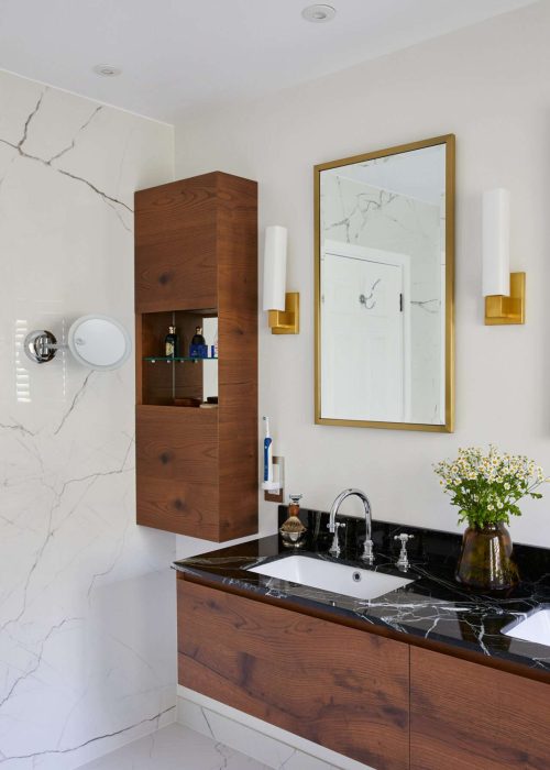 Modern bathroom features a marble sink, wooden cabinetry, large mirror, and elegant lighting. Flowers in a vase add a touch of nature.